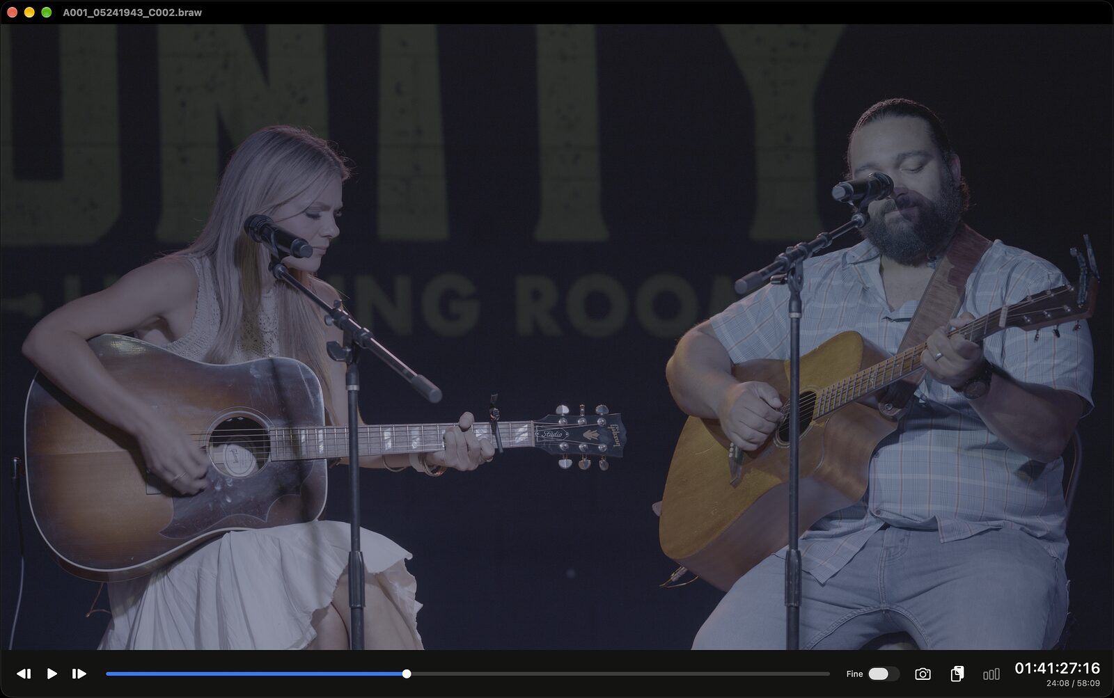 A Blackmagic RAW clip of two musicians on stage, played flat with no look applied: low contrast and washed-out colour